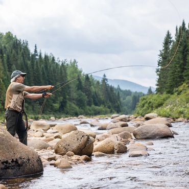 Man fishing with a rod by a rocky river surrounded by forest.