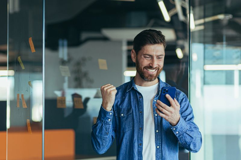 Excited happy business man with a beard, a professional winner, celebrating success by reading good news on a smartphone, feeling excited about mobile messaging