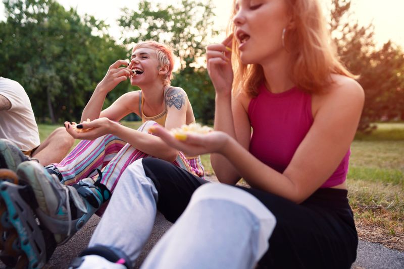 A small group of smiling Caucasian skaters sitting on the ground in a park, talking and eating popcorn together.