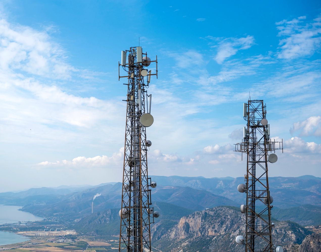 Two communication towers stand tall against a mountainous landscape under a blue sky.
