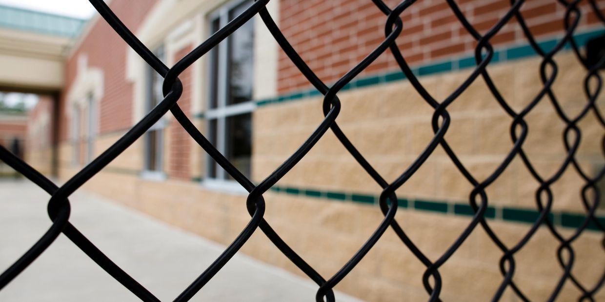 Close-up of a black chain-link fence with a brick building in the background.