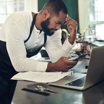 Man in apron looking stressed while checking phone at desk with laptop and papers.