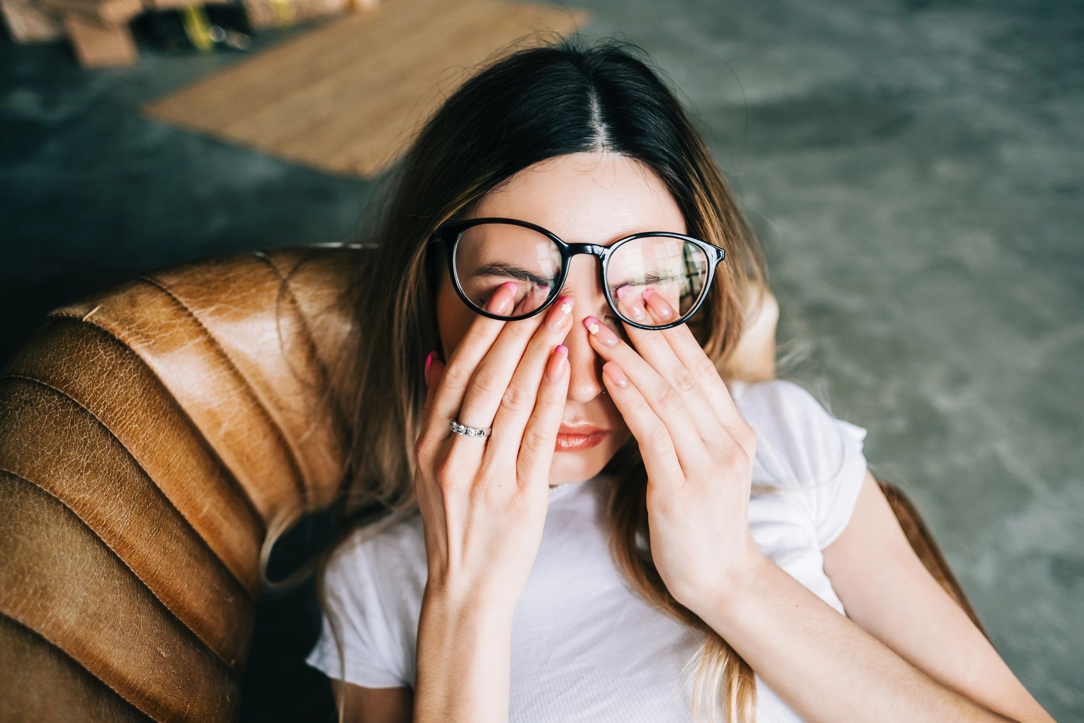Young woman in glasses rubbing her eyes, sitting on a leather chair.