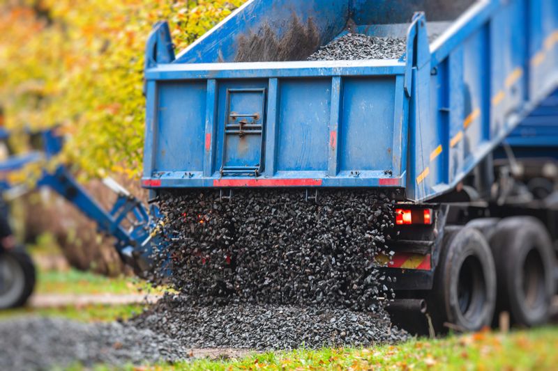 Dump truck, tractor and bulldozer unloading gravel, road metal, rubble and crushed stone cement material during landscaping improvement and new pedestrian walk path road construction site