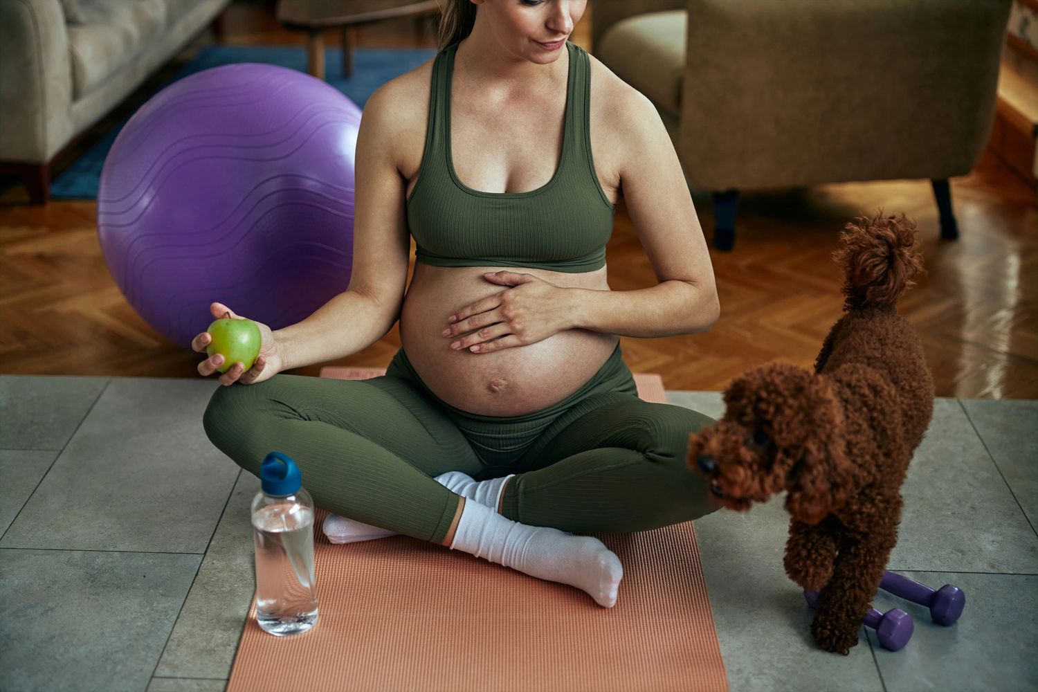 Pregnant woman in workout clothes holding an apple, sitting on a yoga mat with her dog nearby.