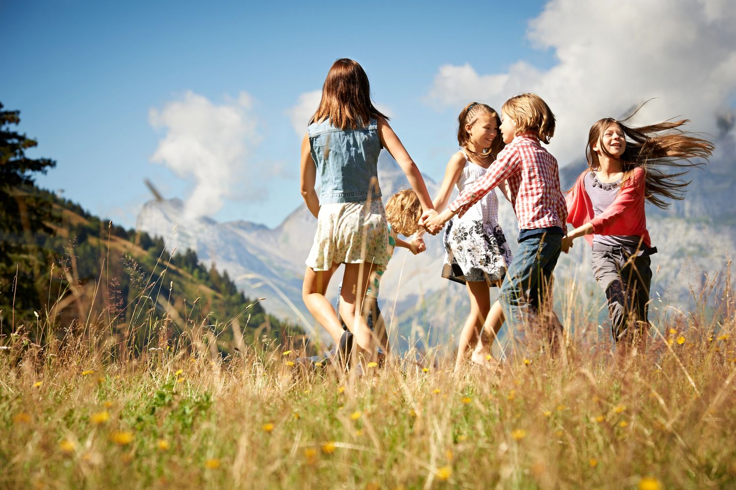 Children holding hands and playing in a sunny meadow with mountains in the background.