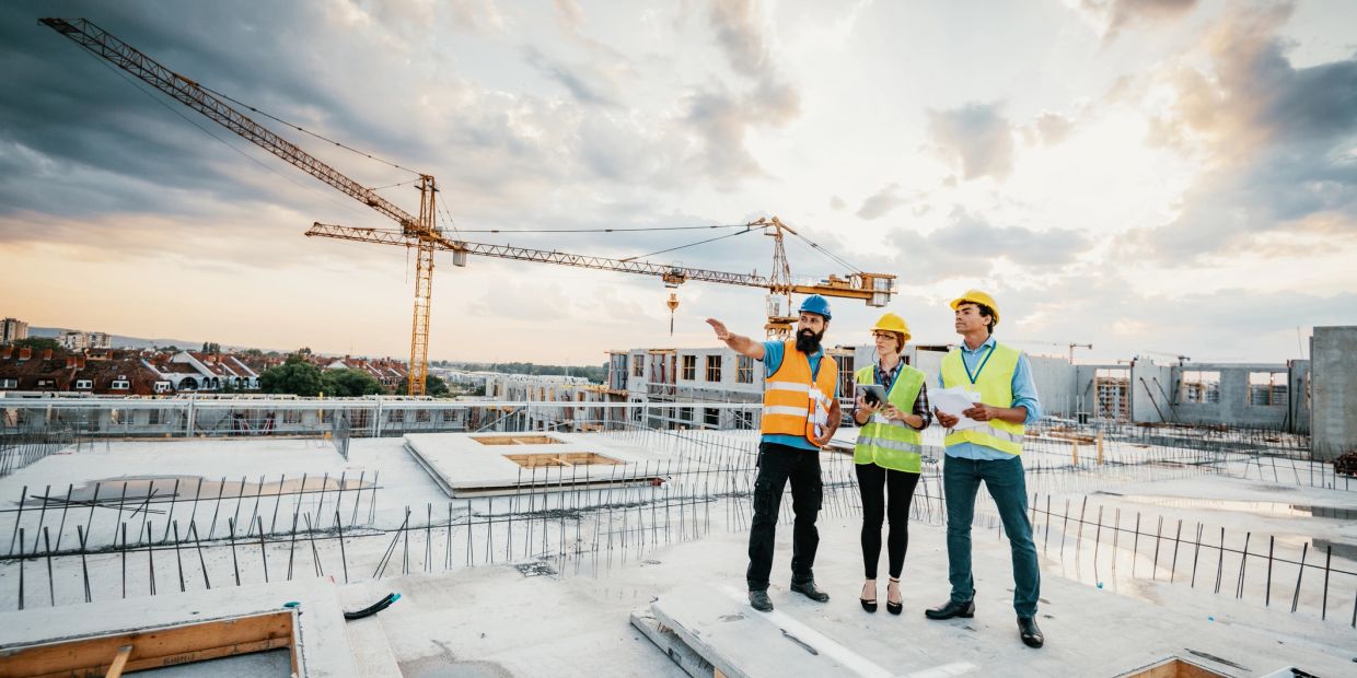 Three construction workers inspect a building site with cranes in the background.