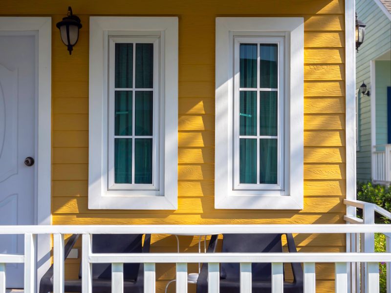 Two white house windows with glass, green curtain decoration on the yellow wooden resident near the front door and terrace, view from outdoor.