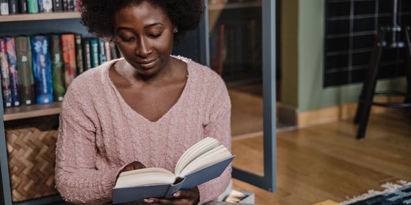Black women with pink V-Neck knit sweater reads a book in a home library. Spark DHE