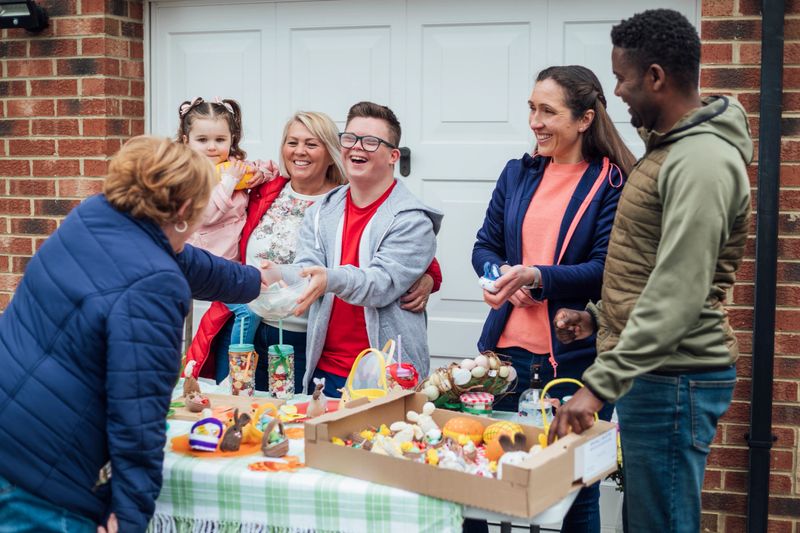 Family having an Easter yard sale in their garden in the North East of England. They have hand made easter crafts on a table and chocolate snacks. One of the sellers has down syndrome and is laughing while collecting money from a customer in a bowl.