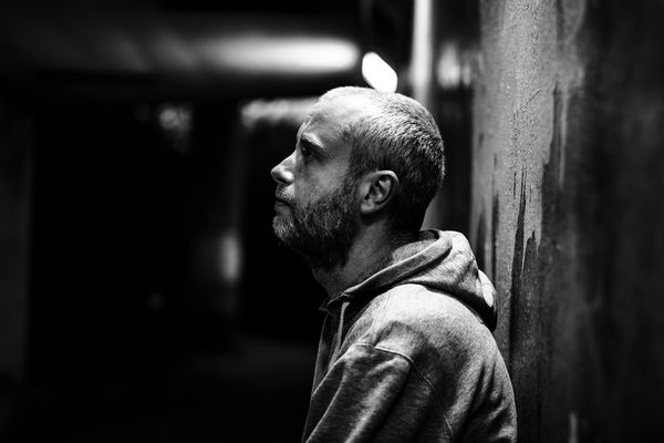 A man with a beard in a hoodie leans against a wall, looking up thoughtfully.