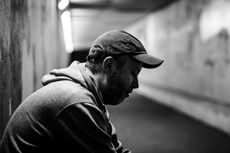 Monochrome toned image depicting a homeless man in his 30s alone in a cold, dark subway tunnel beneath the city. He has a sad, desperate expression on his face, and the man wears a grey hooded top. Focus on the man while the illuminated tunnel recedes into the distance. Room for copy space.