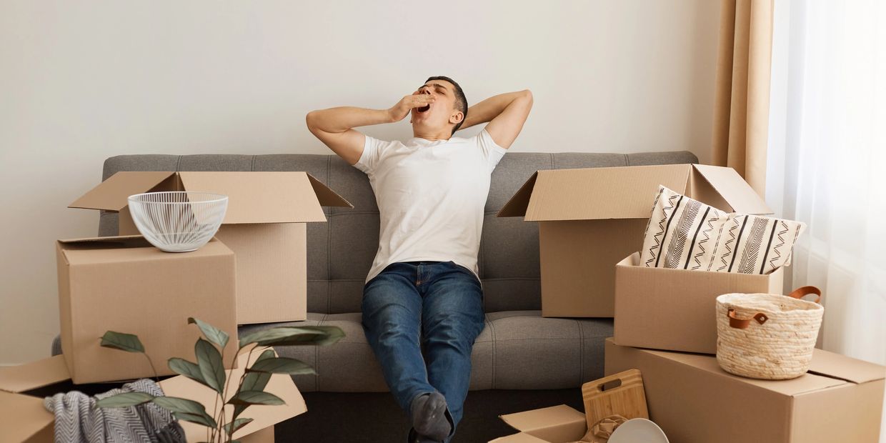 A man yawning while sitting on a couch surrounded by moving boxes.