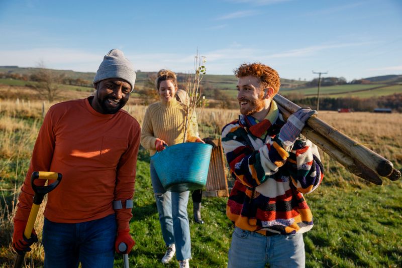 A multiracial group of people wearing warm casual clothing on a sunny cold winters day. Carrying posts and a young tree, friends walk with a man using a crutch. They are working on a community farm, planting trees and performing other sustainable and environmentally friendly tasks.