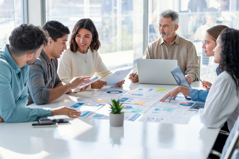 Multi racial group of people working with Paperwork on a board room table at a business presentation or seminar. The documents have financial or marketing figures, graphs and charts on them. People are pointing to different documents. They are happy and smiling. There are laptops and digital tablets on the table