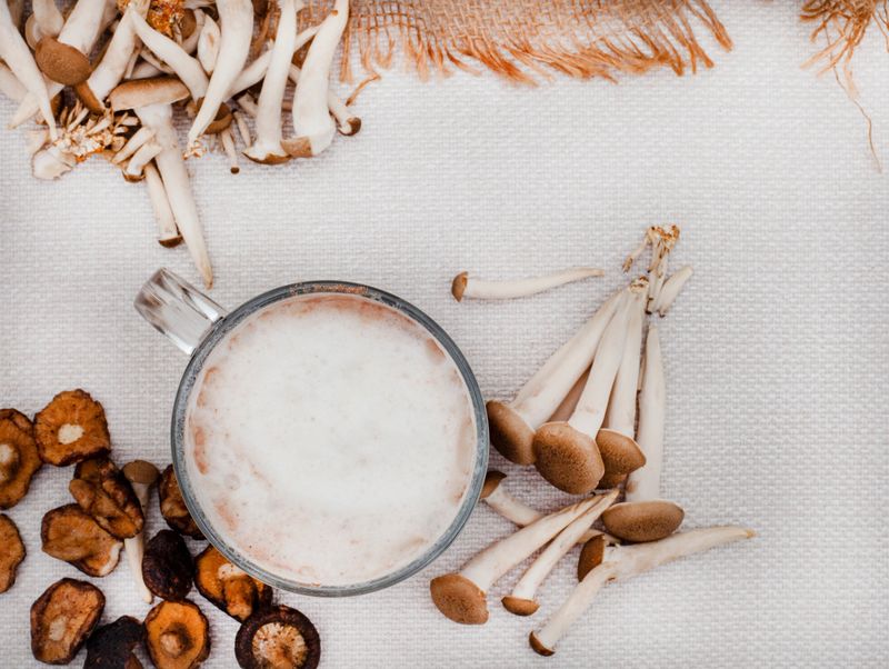 flat lay of mushroom latte in glass mug surrounded with various mushrooms on rustic surface