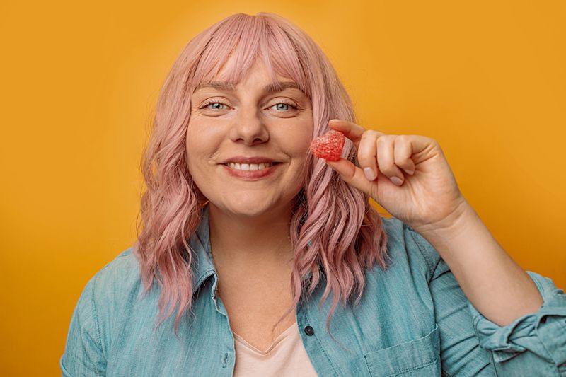 Closeup of a happy woman holding colorful red love shape jelly gummy in hand isolated over yelow wall background. Sweets, confectionery and food concept