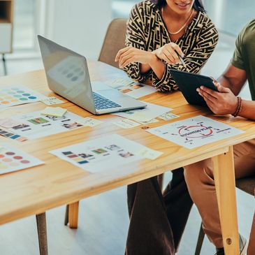 Two creative professionals collaborating at a table with laptop, ipad and colorful printouts.