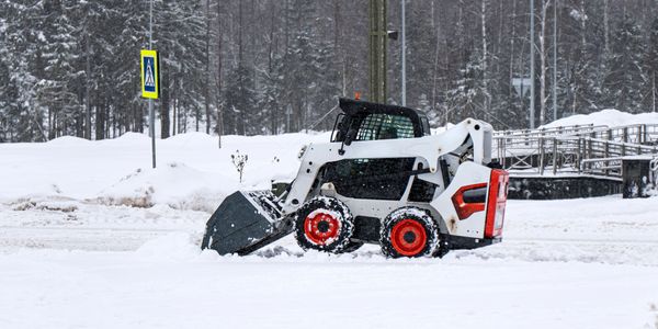 A skid-steer loader clearing snow in a snowy area near trees.