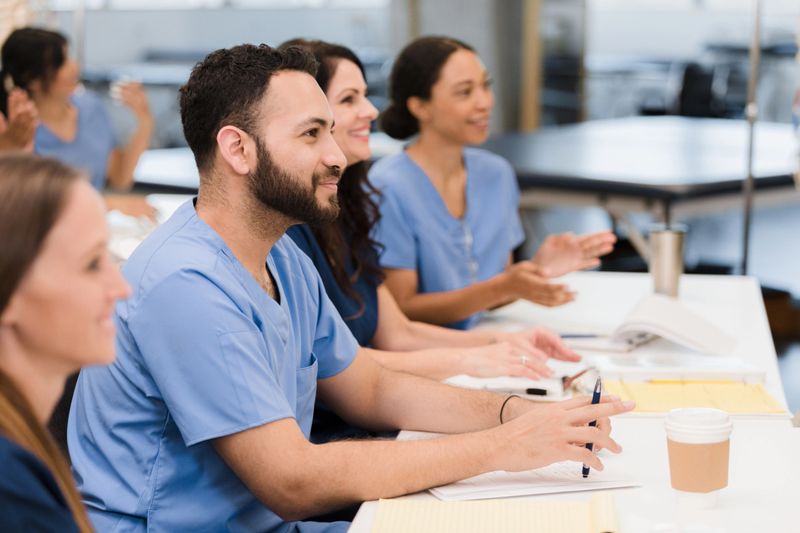 A multi-ethnic group of medical students sit in a row at a table.  They are all listening attentively to a lecture by an unseen professor.