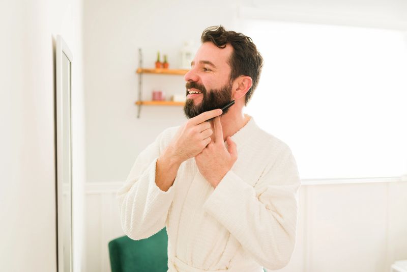 Cheerful young man feeling happy with beard while combing and looking in the mirror before going out