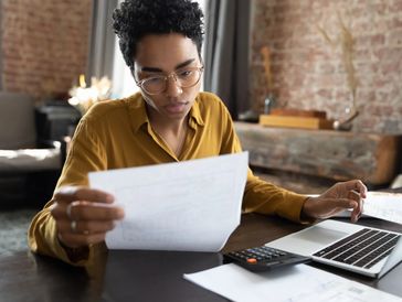 Person in glasses reviewing documents with a laptop and calculator on the table.