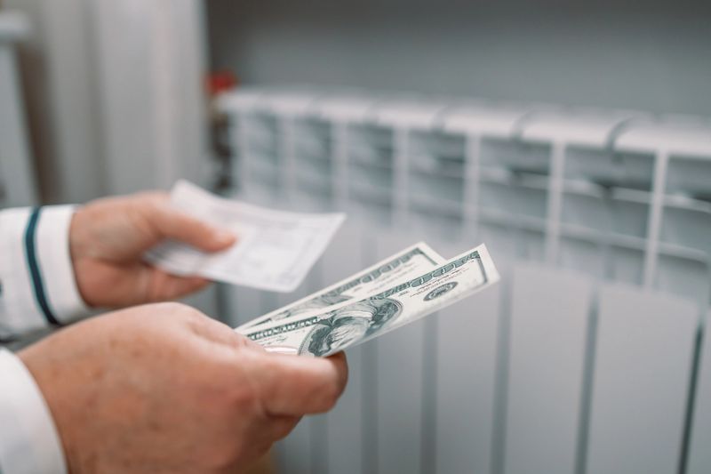 Paying bills. Dollar bills in female hands near heating radiator battery at home. Expensive heating costs.