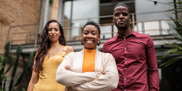 Three confident friends standing outdoors in front of a building, smiling at the camera.