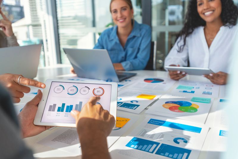 Group of people meeting with technology and paperwork. Close up of digital tablet with modern chart data on the screen. The documents have financial or marketing figures, graphs and charts on them. Multi ethnic group including Caucasian and African American
