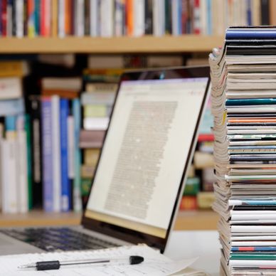 Stack of magazines beside a laptop with bookshelves in the background.