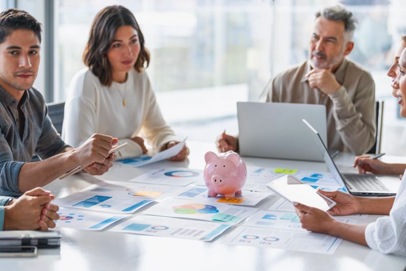 Multi racial group of Business people with a piggy bank. There are people of different ethnic groups. All are dressed in business casual clothing.Paperwork on a board room table at a business presentation or seminar. The documents have financial or marketing figures, graphs and charts on them. There are laptops and digital tablets on the table