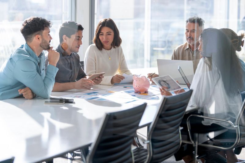 Multi racial group of Business people with a piggy bank. There are people of different ethnic groups. Paperwork on a board room table at a business presentation or seminar. The documents have financial or marketing figures, graphs and charts on them. There are laptops and digital tablets on the table