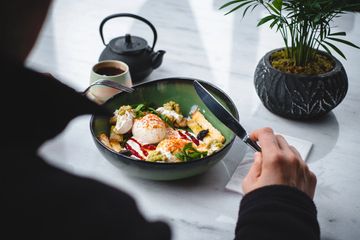 Person about to eat a colorful bowl of food with a teapot and plant nearby.