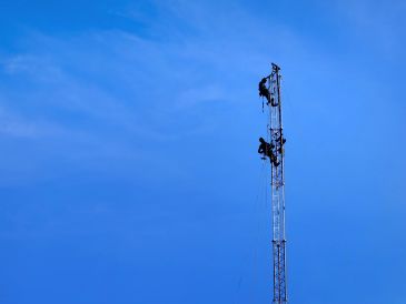 Dois trabalhadores sobem uma torre alta contra céu azul.