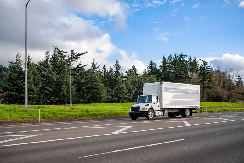 Powerful classic white big rig semi truck tractor transporting commercial cargo in long box trailer for local deliveries running on the highway entrance for the next delivery