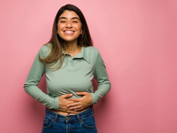 Young woman smiling and holding her stomach happily against a pink background.