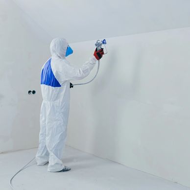 Person in protective gear spray painting a white wall indoors.