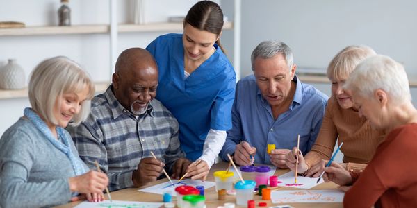 A group of elderly people painting together with a caregiver's assistance.