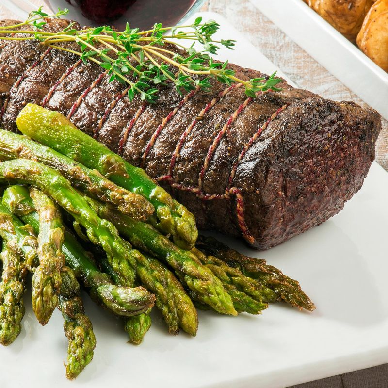 Whole beef tenderloin cooked in the oven, tied with a rope in a white porcelain plate, and the asparagus next to it.