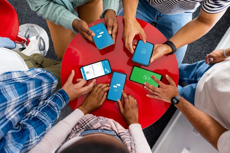 Direct above shot of student friends sitting together around a table using their phones to bank transfer money to one another. The app is open on all of their smart phone device screens.  They are studying at university in the North East of England.