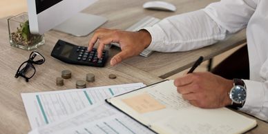 Person using calculator and writing notes with coins and documents on desk.