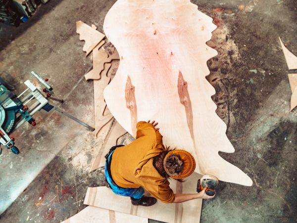 Person sanding a large wooden wing-shaped piece in a workshop.