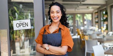 Smiling woman in apron stands by an open sign at a cafe entrance.