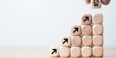 Wooden blocks stacked in a staircase with arrows pointing up and a hand placing a quality badge block on top.