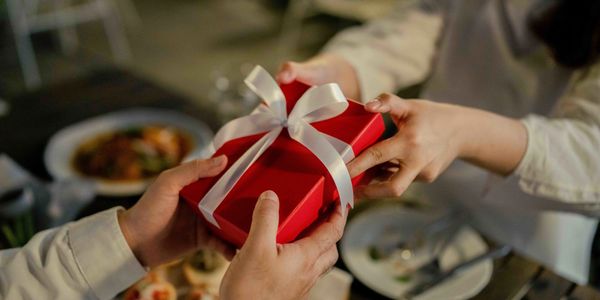A gourmet gift is being handed to a loved one in a red box with a white ribbon tied around it.