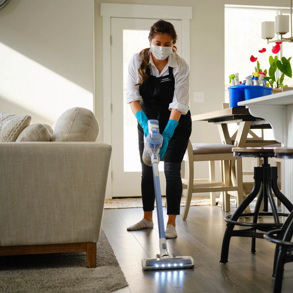 A masked woman vacuuming a living room floor with gloves and apron.