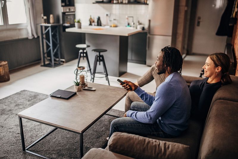 Two people, black male and his white girlfriend sitting on sofa in living room and watching television together.