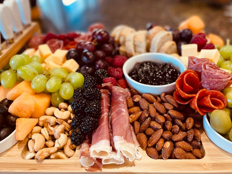 Surface level tabletop view charcuterie board fresh fruit cured meats cheeses nuts crackers bread, French-inspired branch of cooking, delicious healthy appetizers on bamboo plater or board.