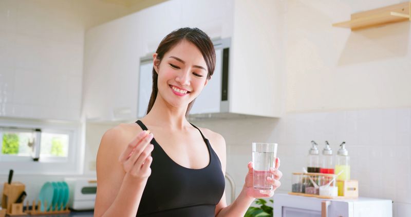 asian fitness woman taking nutrient supplement or medicine by water in kitchen at home