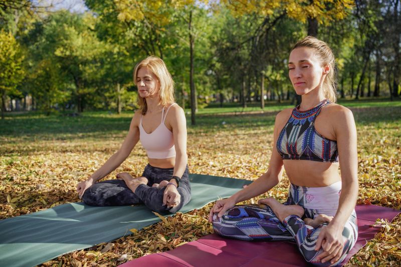 Two women sitting in lotus position on yoga mats in the park, keeping eyes closed in silence, relaxation technique for stress relief